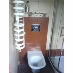 Wnętrze łazienki z brązowymi płytkami, wiszącym grzejnikiem drabinkowym, toaletą i fragmentem kabiny prysznicowej; na półce nad toaletą stoi butelka płynu.